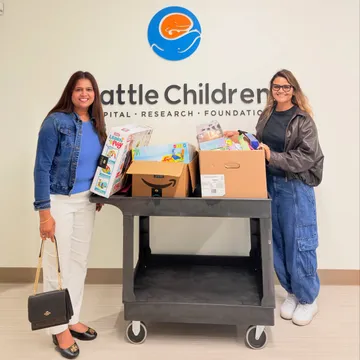 Two Spark WA volunteers standing beside a cart loaded with donated toy and school-supply boxes, delivered to a Seattle-area children's hospital